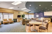 Open senior living common/dining area with round tables and chairs, a lounge of armchairs by large windows, a wall-mounted TV, and a kitchenette/bar.