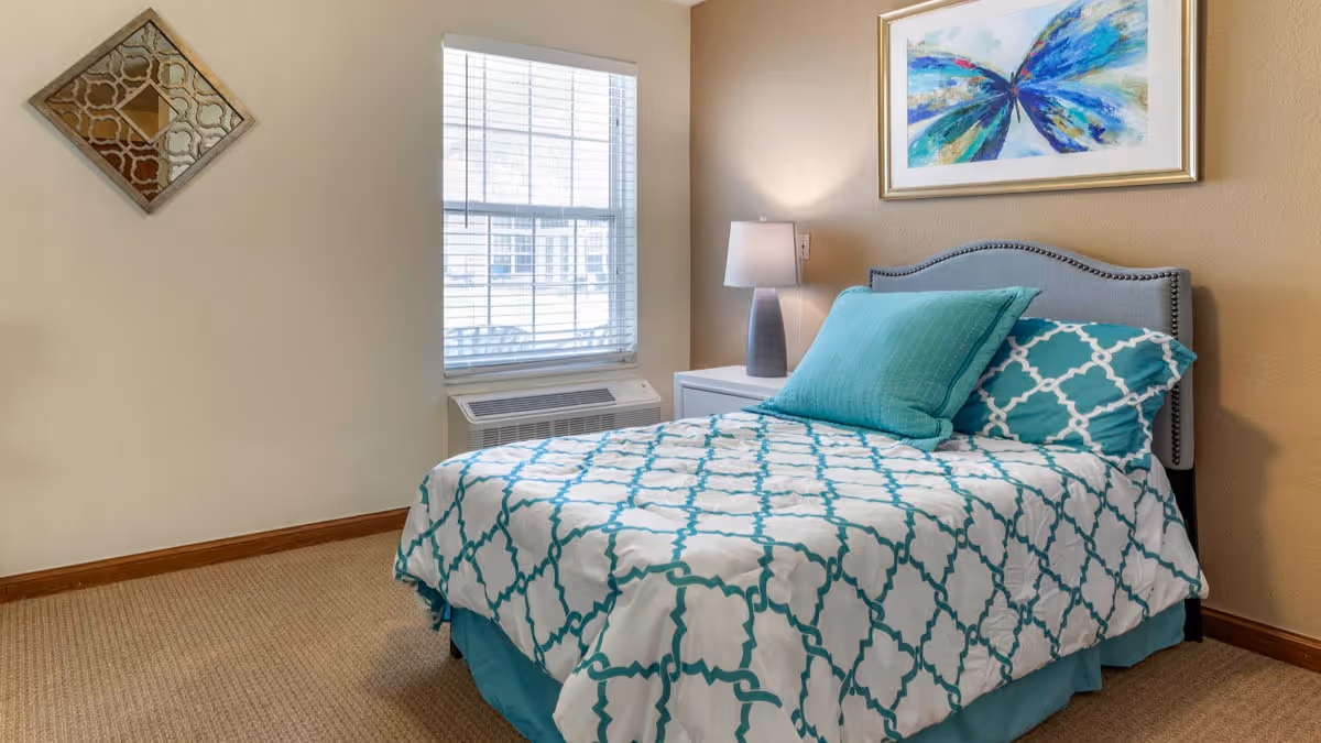 A neatly made single bed with a white and teal patterned comforter and matching pillows in a small bedroom. There is a window with blinds, a white nightstand with a lamp, a decorative wall mirror, and a colorful abstract painting above the bed.