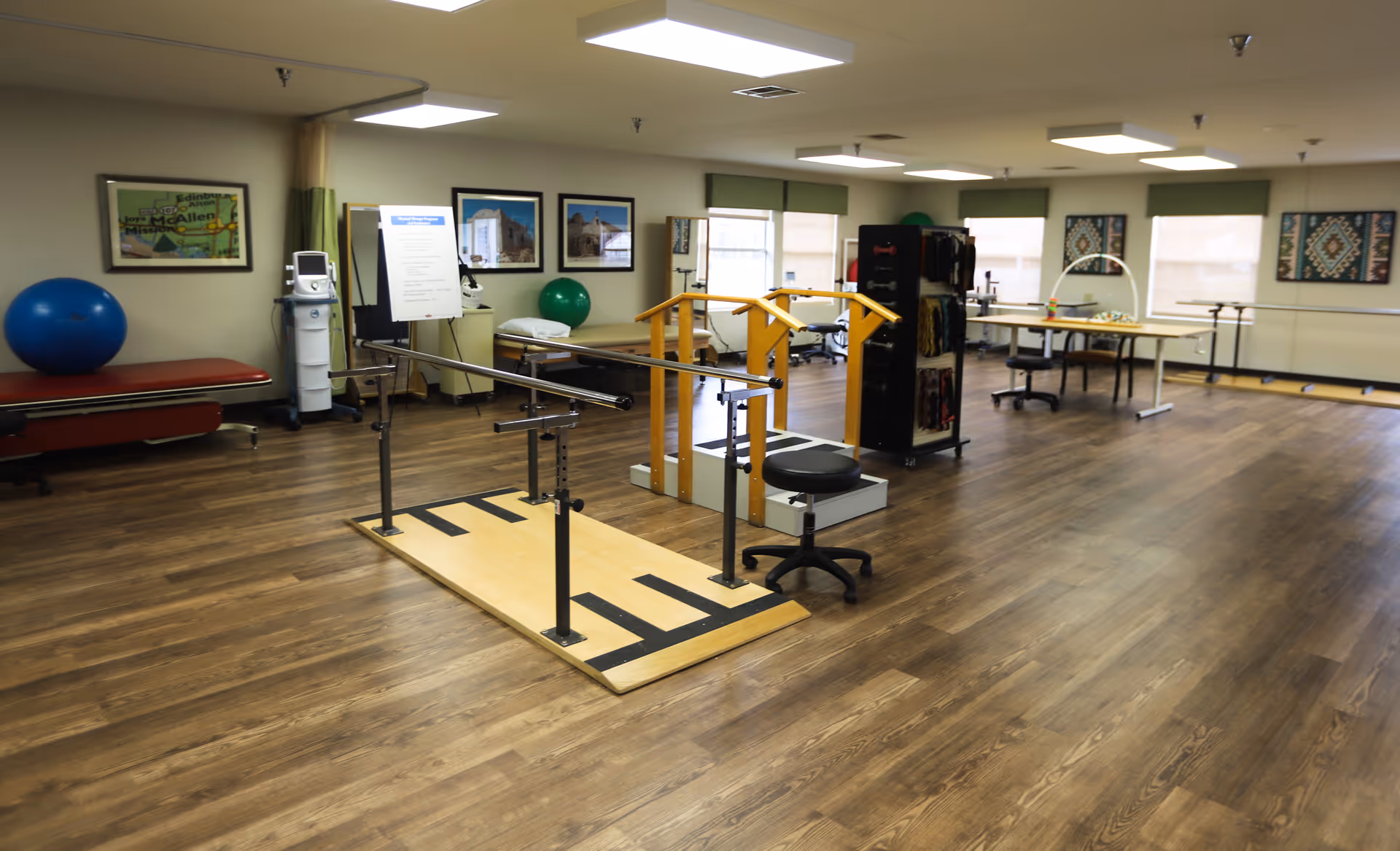 A spacious rehabilitation room with wooden flooring, parallel bars for walking exercises, exercise balls, therapy equipment, chairs, and tables. The room has several windows with green blinds and framed pictures on the walls.