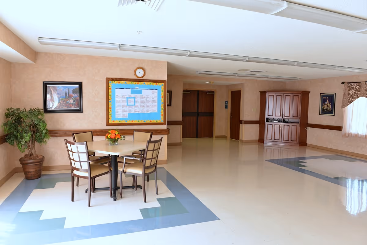 A spacious common area in a senior living facility with a small square table surrounded by four chairs. The floor has a decorative pattern with blue and white tiles. There is a potted plant in the corner, framed artwork on the walls, a bulletin board with notices, and a wooden cabinet. The room is well-lit with natural light coming through a window with a valance curtain.