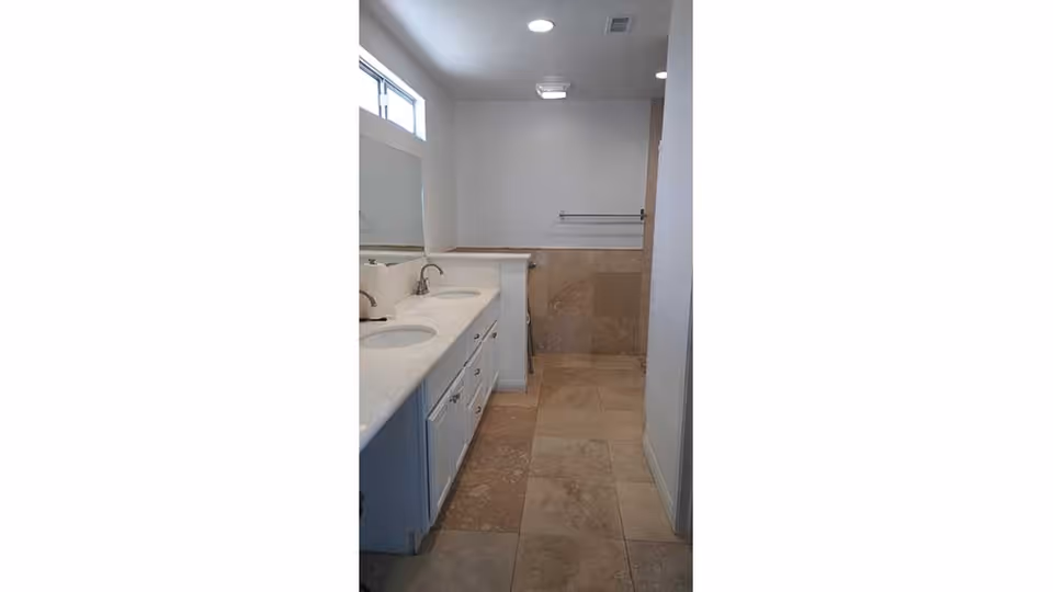 A bathroom with beige tiled floor and walls, featuring a white double sink vanity with silver faucets and a large mirror above it. There is a window near the ceiling allowing natural light in, and a towel rack mounted on the wall opposite the sinks.