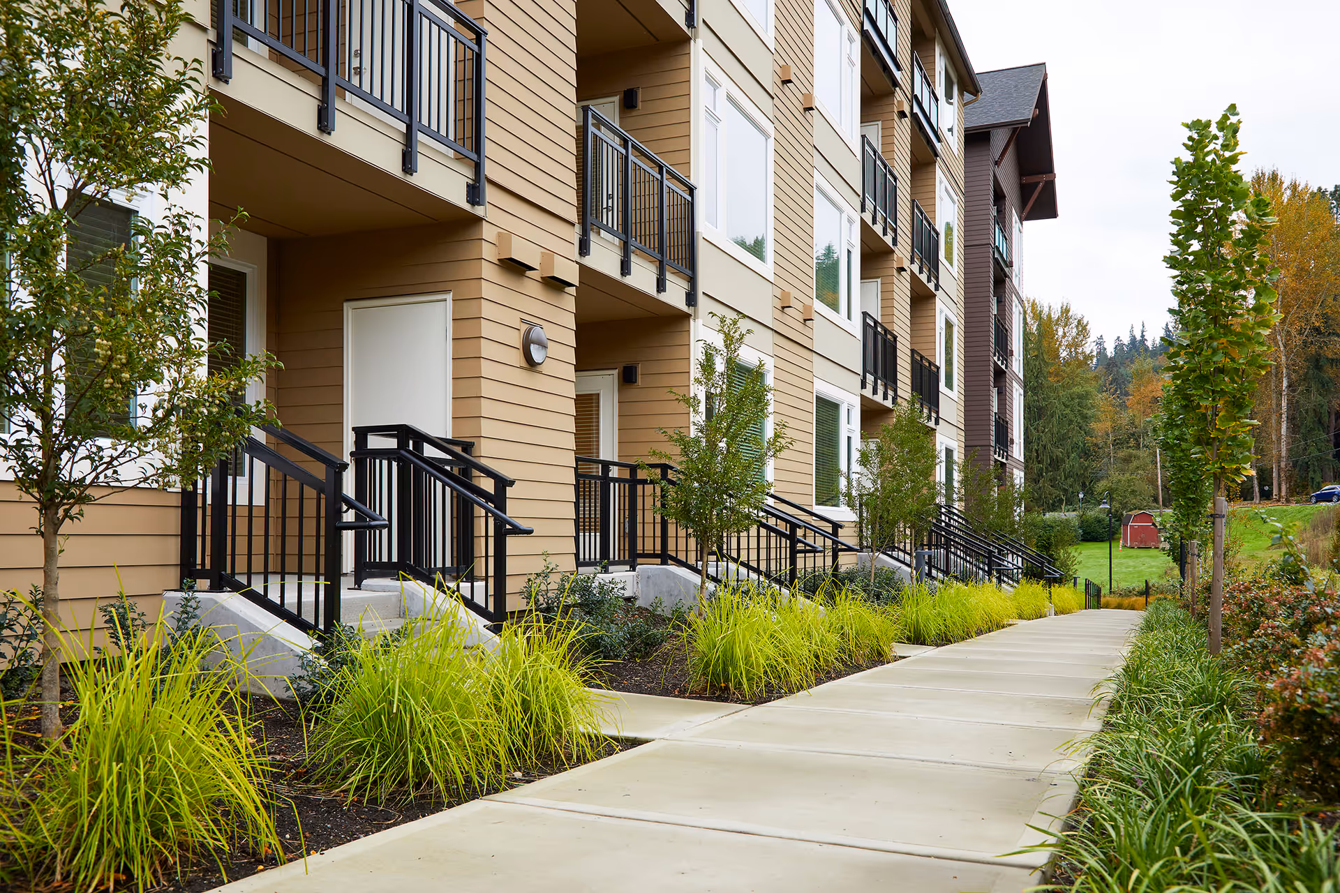 Sidewalk and landscaped path leading past entrances, stairs, and balconies of a modern multi-story residential building.