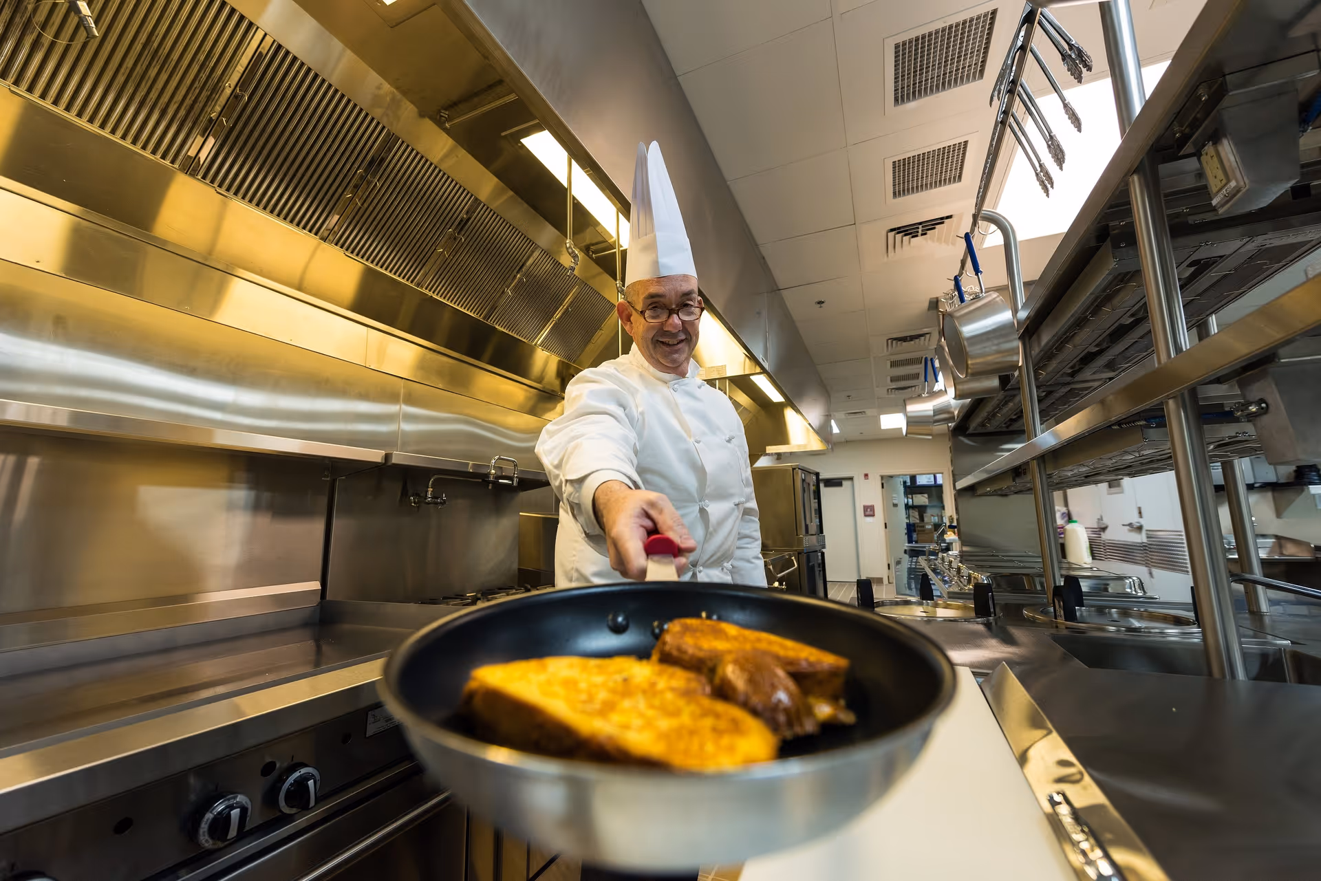 A chef wearing a white uniform and tall white chef hat is smiling and holding a frying pan with two pieces of cooked food, possibly French toast, in a professional stainless steel kitchen.
