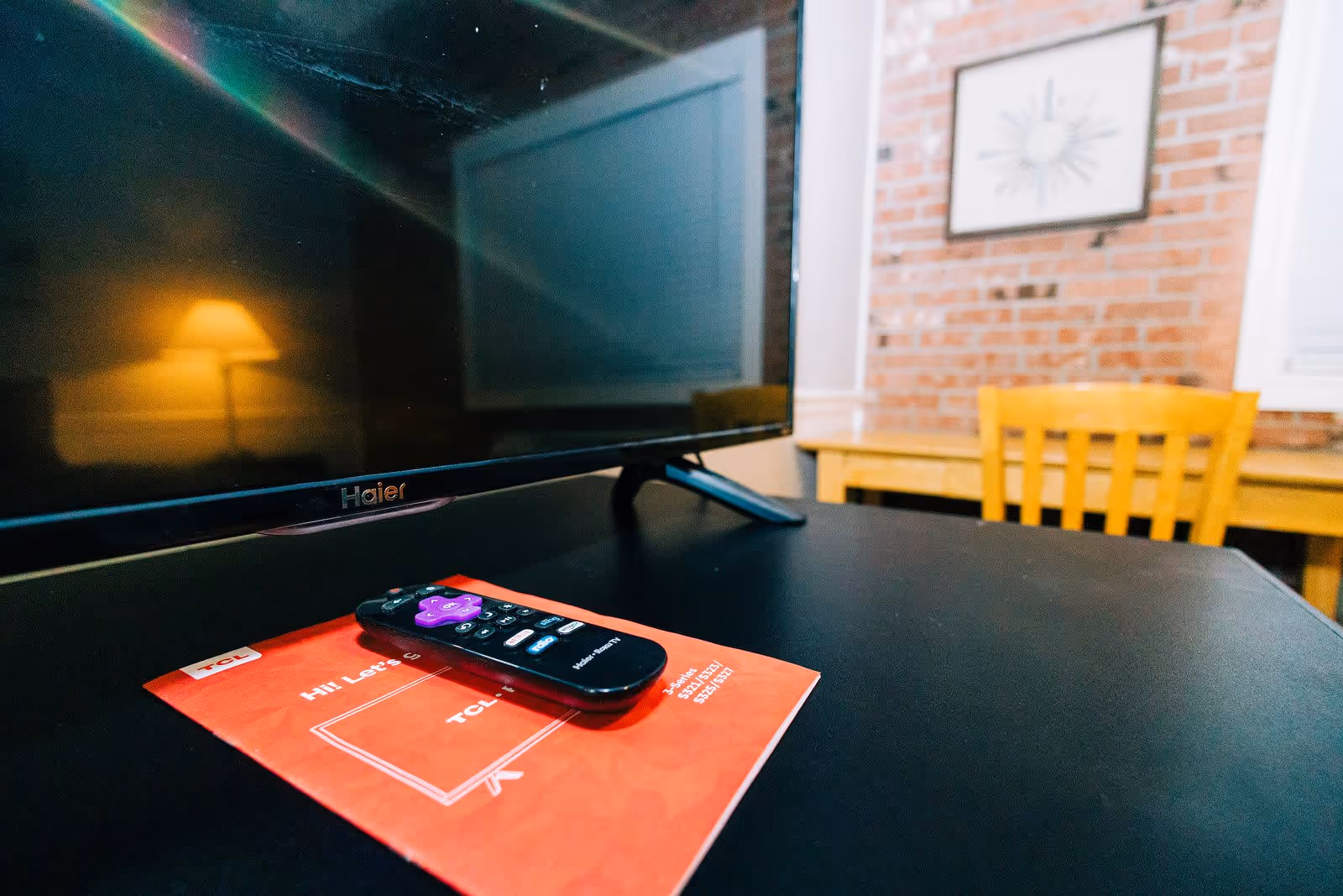 Close-up view of a black Haier television on a black table with a remote control and a red instruction manual placed in front of it. In the background, there is a wooden chair, a wooden table, a brick wall, and a framed picture hanging on the wall.