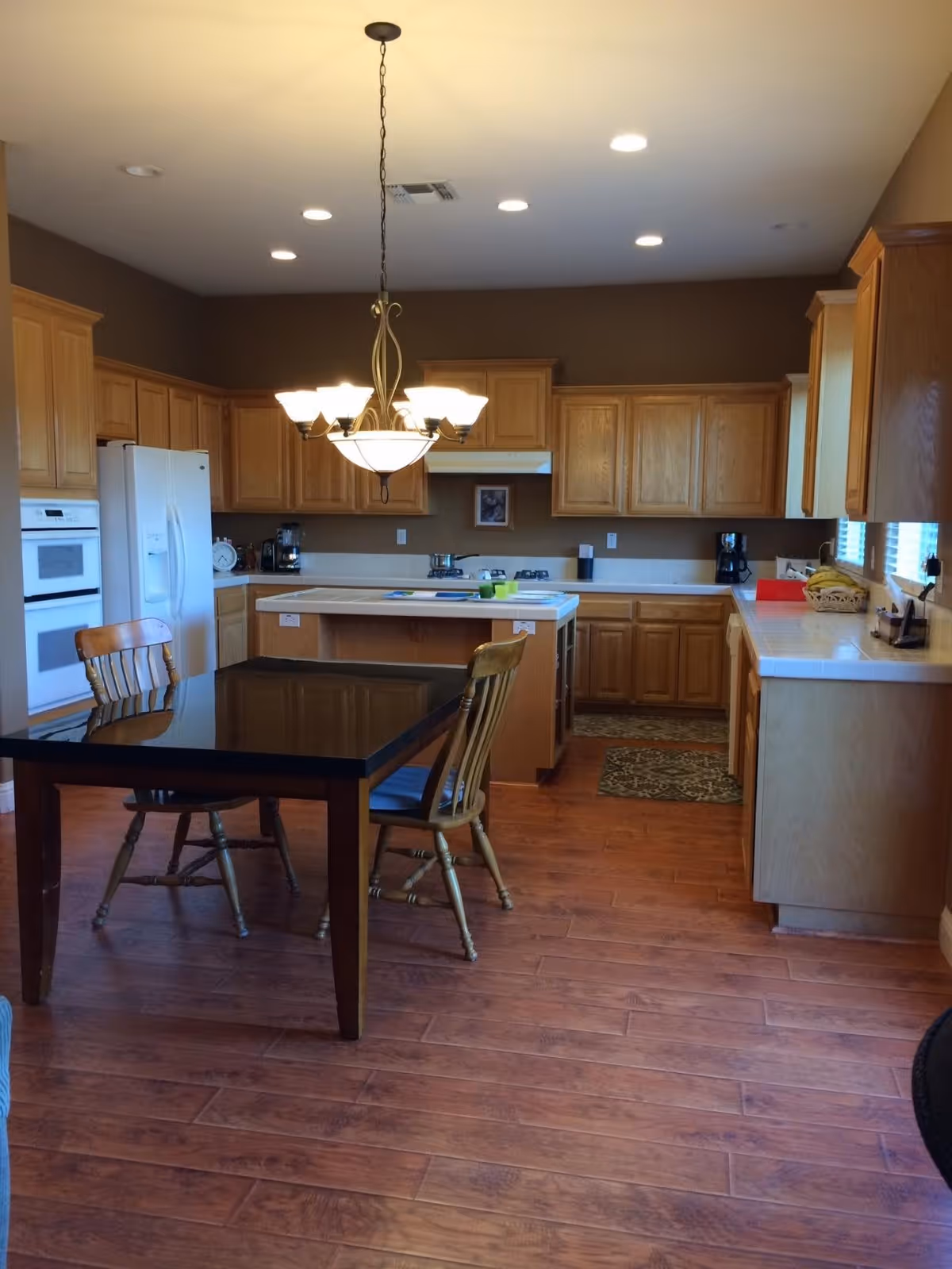 A spacious kitchen with wooden cabinets, a white refrigerator, built-in oven, and a central island. There is a black dining table with two wooden chairs in the foreground. The floor is wooden, and a chandelier with multiple lights hangs from the ceiling.