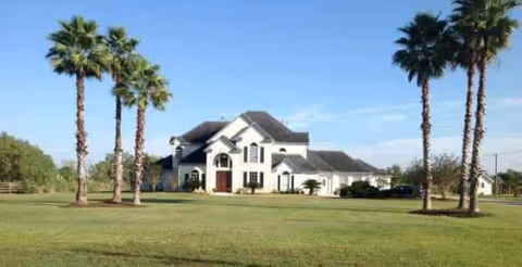 Large white assisted living facility front with arches on a grassy lawn and palm trees.