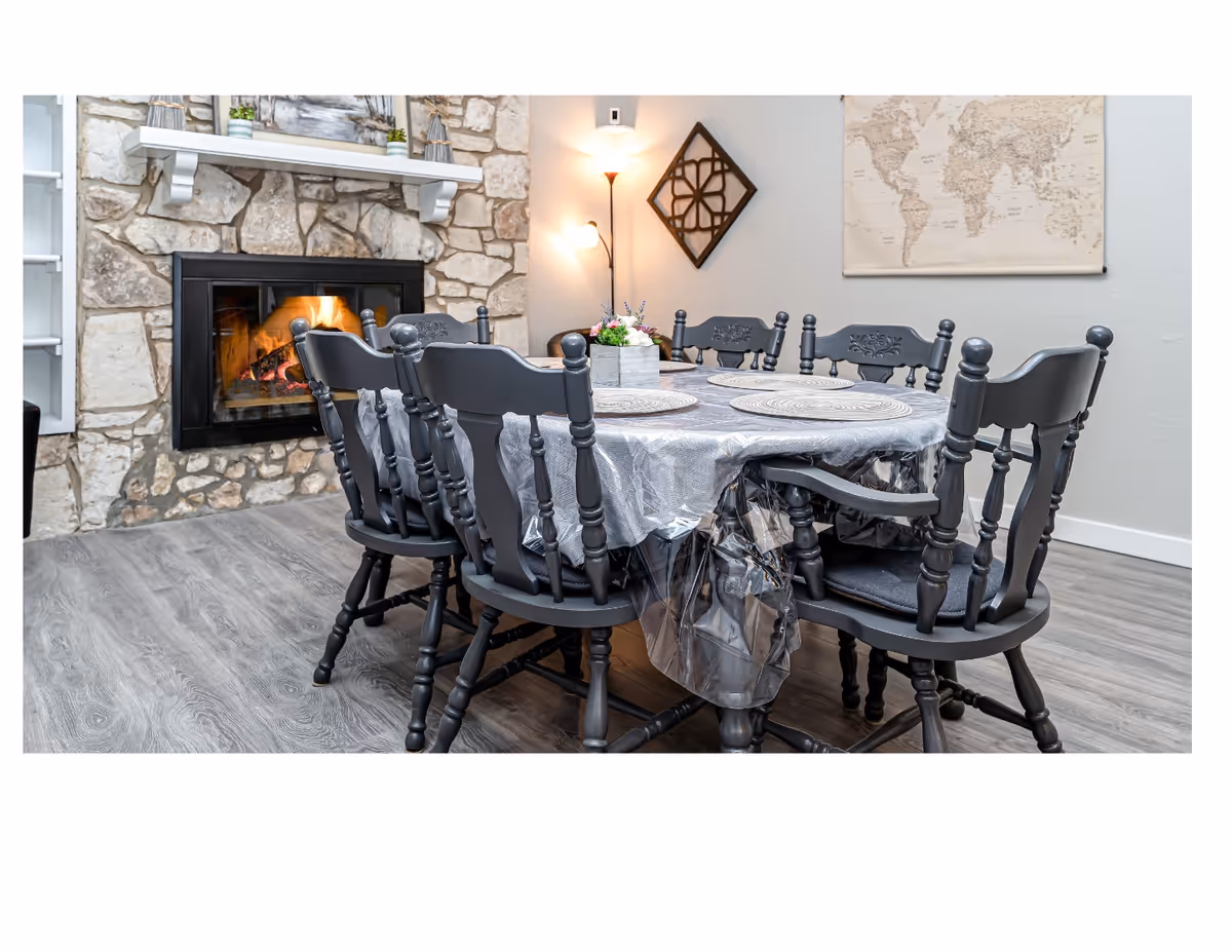 A dining area with a round table covered by a plastic tablecloth and six dark wooden chairs around it. The table is set with four round placemats and a small floral centerpiece. In the background, there is a stone fireplace with a fire burning inside, a floor lamp, a decorative wall piece, and a world map hanging on the wall. The floor is covered with wood-like vinyl flooring.