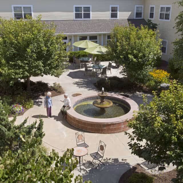 Outdoor courtyard area with a circular brick fountain in the center, surrounded by trees, shrubs, and patio furniture including tables with umbrellas and chairs. Two elderly people are standing and sitting near the fountain, engaged in conversation. The courtyard is adjacent to a light-colored building with multiple windows.