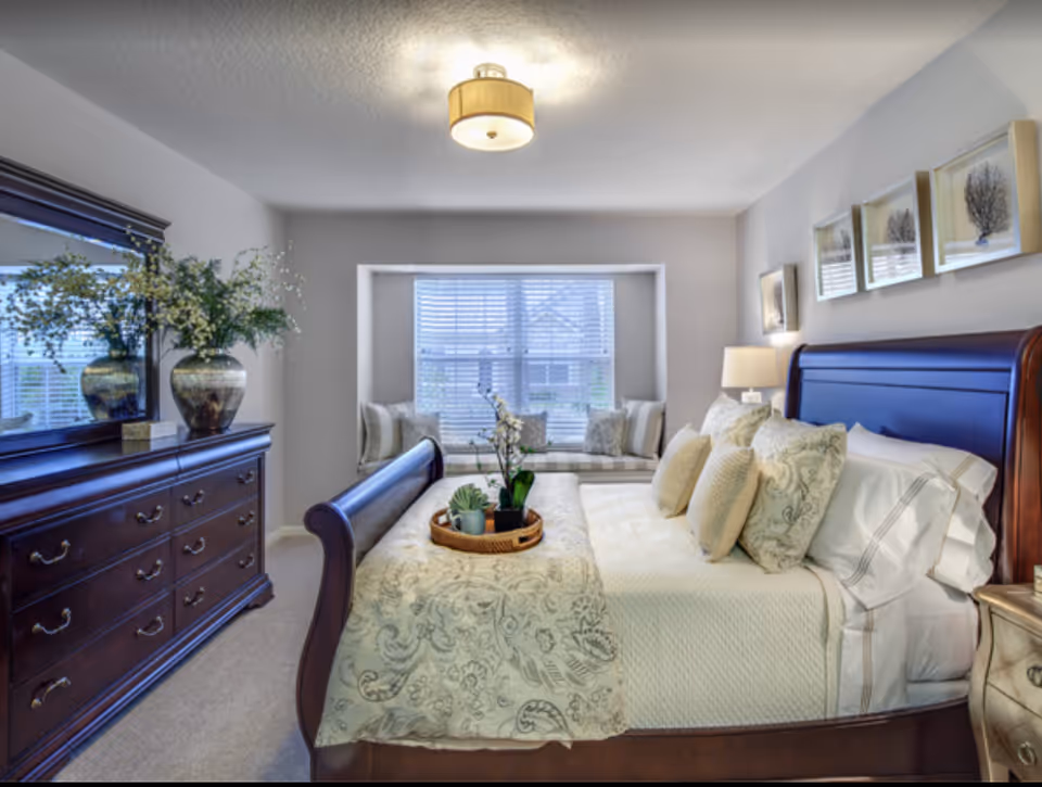 A well-lit bedroom featuring a large wooden bed with multiple pillows and a patterned bedspread. A wooden dresser with a large mirror and decorative vases is on the left side. A window seat with cushions is positioned under a large window with blinds, and framed artwork hangs above the bed.