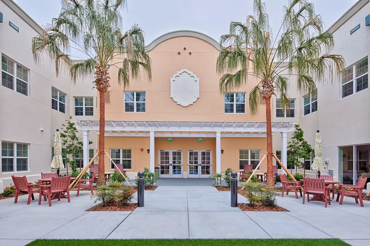 Outdoor courtyard area at Lexington Place featuring palm trees, red wooden chairs and tables, umbrellas, and a beige building with multiple windows and double glass doors under a white pergola.