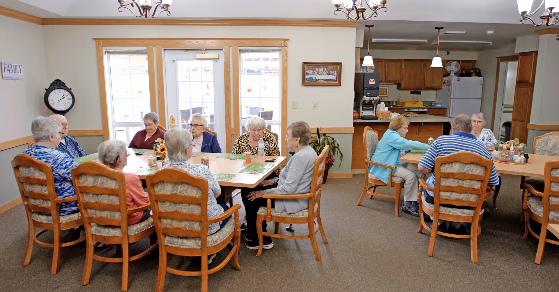 Several elderly residents seated around two tables in a communal dining area with windows and a kitchenette in the background.