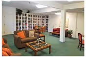A spacious living room area with green carpet, featuring brown sofas with orange and beige cushions, a glass coffee table, a bookshelf filled with books, and a small dining table with chairs. There are white walls and ceiling with fluorescent lighting.