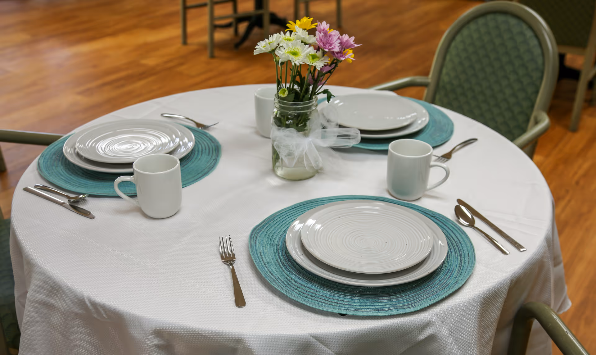 Round dining table set for three with white plates, teal placemats, white mugs, cutlery, and a mason jar of flowers on a white tablecloth.