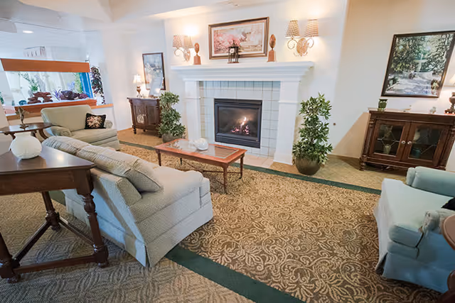 A cozy living room with a lit fireplace in the center, flanked by two potted plants. The room features patterned carpet, a wooden coffee table, two armchairs, and a sofa. There are framed artworks on the walls and decorative lamps on side tables.