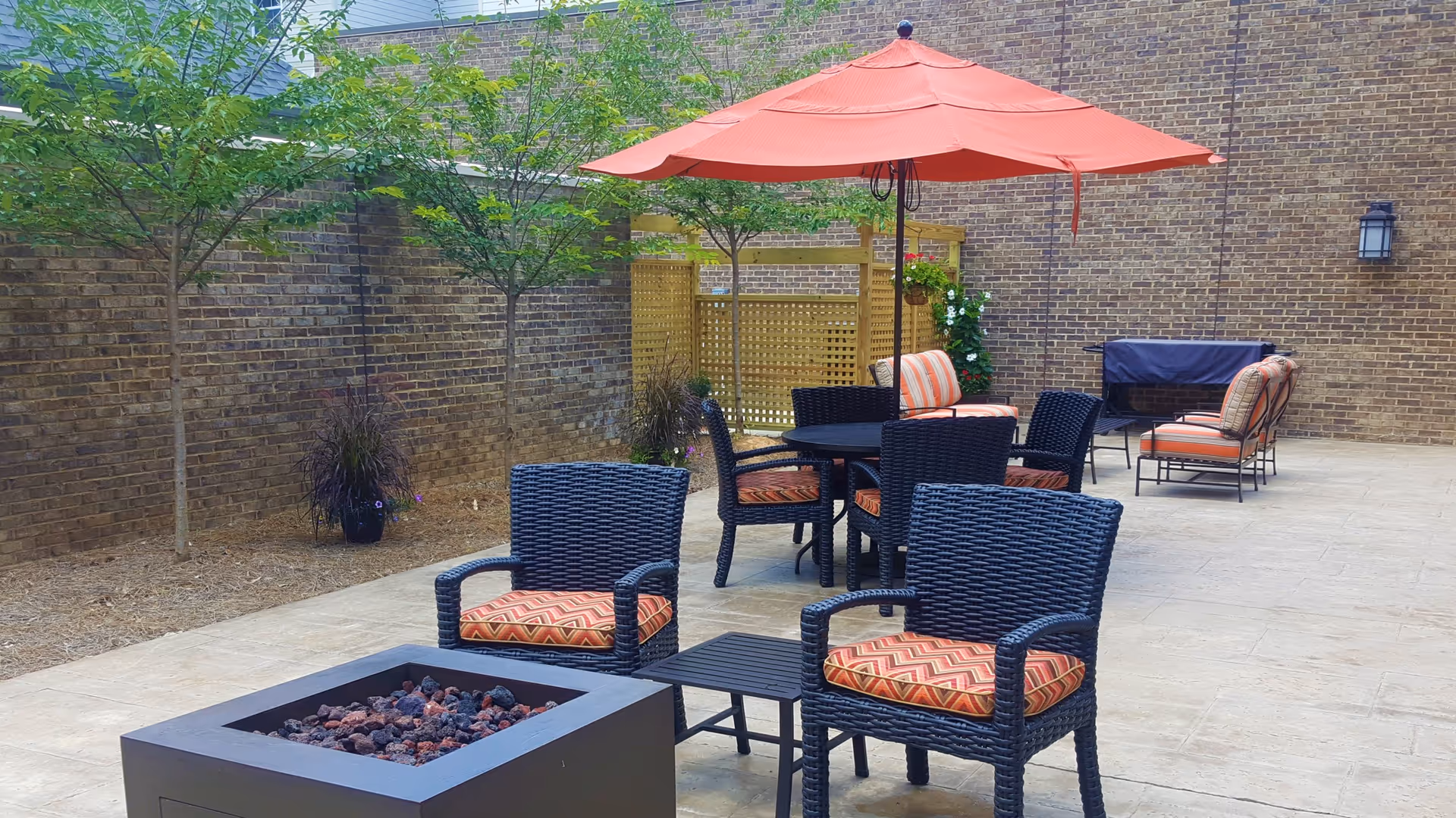 Outdoor patio area with black wicker chairs featuring orange patterned cushions, a square fire pit, a round table with an orange umbrella, additional seating with striped cushions, small trees, and a brick wall background.