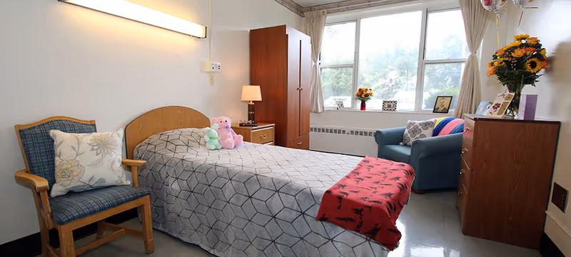 A small, cozy bedroom with a single bed covered in a gray patterned blanket and a red throw at the foot. Two stuffed animals are placed on the bed. To the left of the bed is a wooden chair with a floral cushion. Next to the bed is a wooden nightstand with a lamp. A wooden wardrobe stands against the wall near a large window with beige curtains. In front of the window is a blue armchair with a decorative pillow and a colorful blanket draped over one arm. To the right is a wooden dresser with a vase of sunflowers, framed photos, and balloons. The room has a clean, simple, and homey atmosphere.