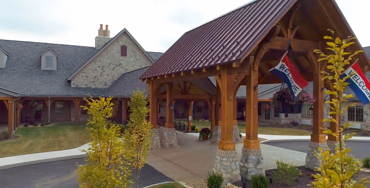 Exterior view of The Inn at Library Way showing a covered entrance with wooden beams and stone pillars, a driveway, landscaped greenery, and a building with a stone facade and multiple windows under a gray roof. Two flags with the word 'WELCOME' hang from the wooden structure.