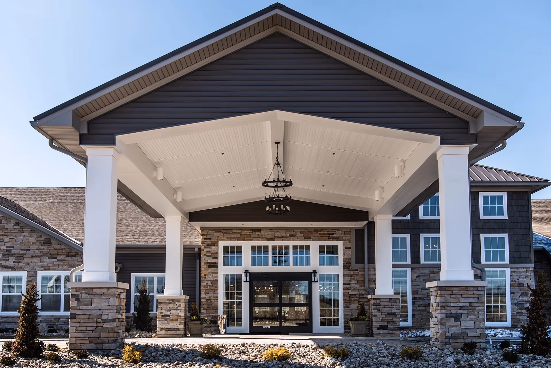 Front exterior view of a senior living facility with a large covered entrance supported by white columns with stone bases, large windows, and a clear blue sky.
