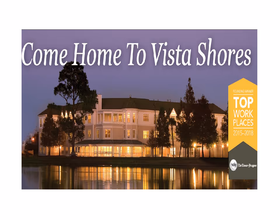 Evening view of a large, well-lit senior living facility building reflected in a calm body of water, with trees in the foreground and the text 'Come Home To Vista Shores' at the top.
