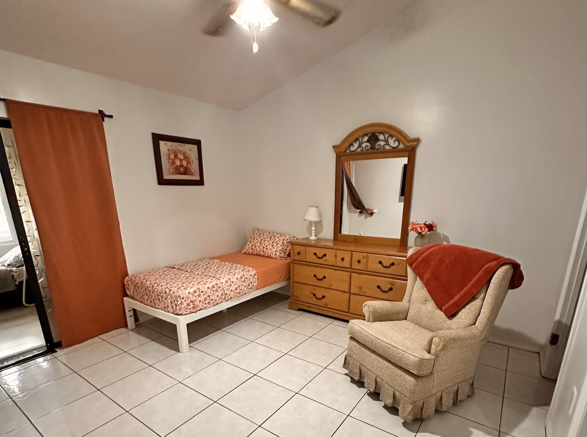 A simple bedroom with a single bed covered in orange and floral bedding, a wooden dresser with a mirror, a small lamp, and a beige armchair with a red blanket draped over it. The room has white tiled floors, white walls, a ceiling fan with a light, and an orange curtain covering a sliding glass door.
