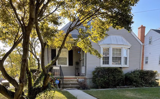 Exterior view of a single-story house with gray siding, a small porch with steps, a bay window, and a brick chimney. The house is surrounded by green bushes and trees with sunlight filtering through the leaves. A concrete walkway leads to the front door.