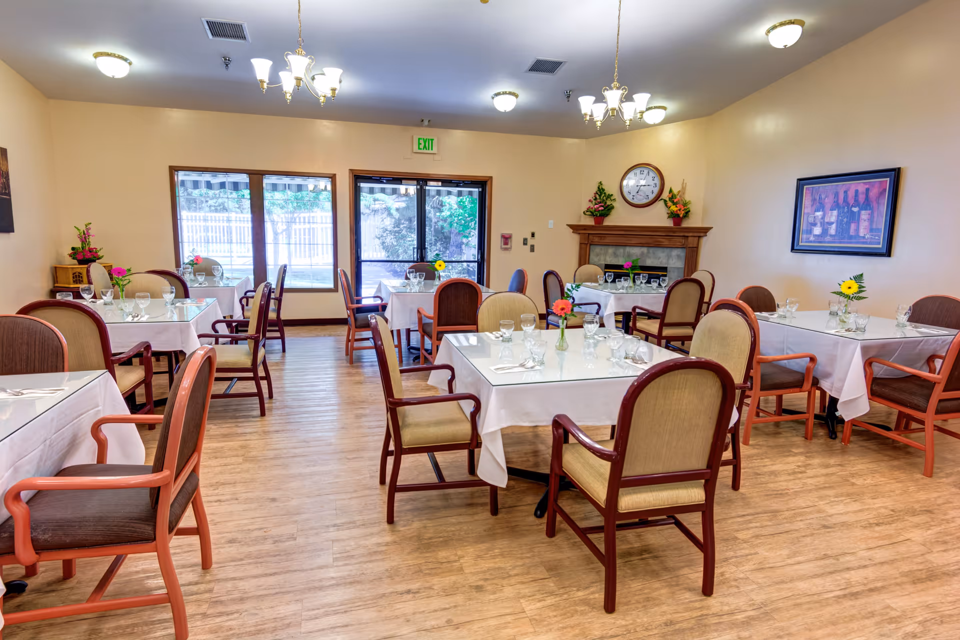 A bright dining room with several tables covered in white tablecloths, each set with glasses, napkins, and small flower vases. The room has wooden chairs with cushioned seats and backs, wood flooring, and large windows letting in natural light. There is a fireplace with a clock and flower arrangements above it, and framed artwork on the walls.