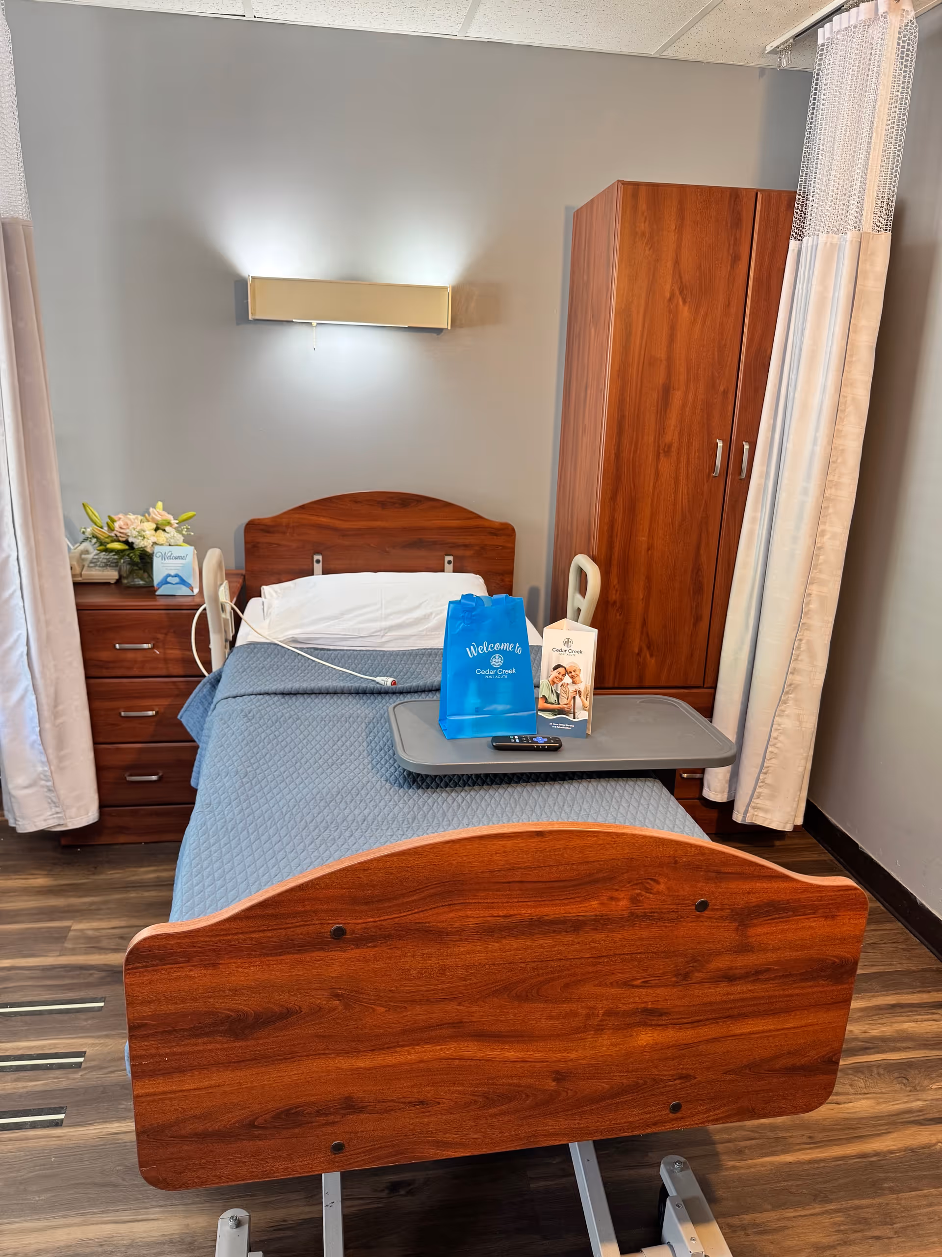 A furnished senior care room with a hospital-style bed, overbed tray holding a welcome bag and brochure, bedside cabinet and wardrobe.