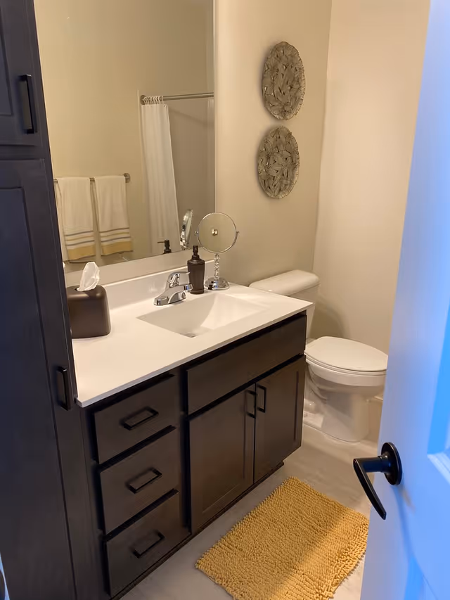 A bathroom with a white countertop sink, dark wood cabinets, a toilet, a yellow bath mat, two decorative wall hangings, and a mirror reflecting a towel rack with two white towels.