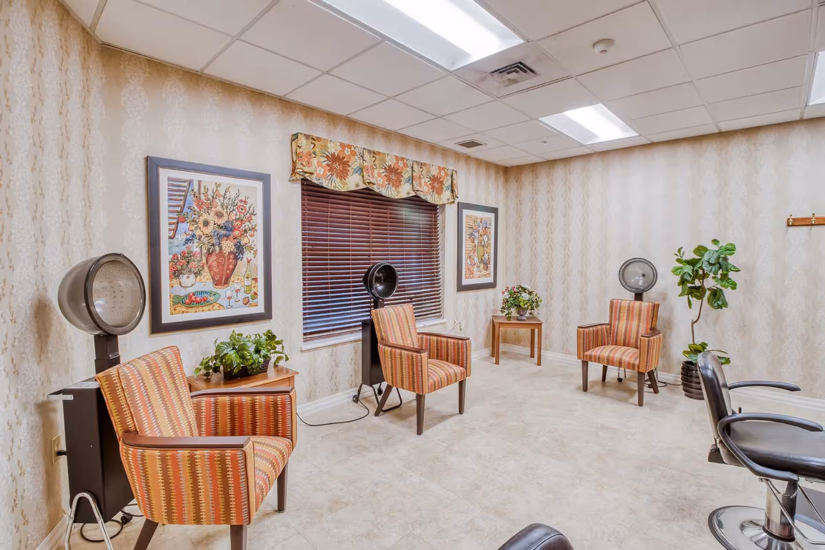 Interior view of a small salon area in a senior living facility with three striped armchairs, two vintage hair dryers, framed floral paintings on the wallpapered walls, a window with blinds and a floral valance, small tables with plants, and a salon chair in the foreground.