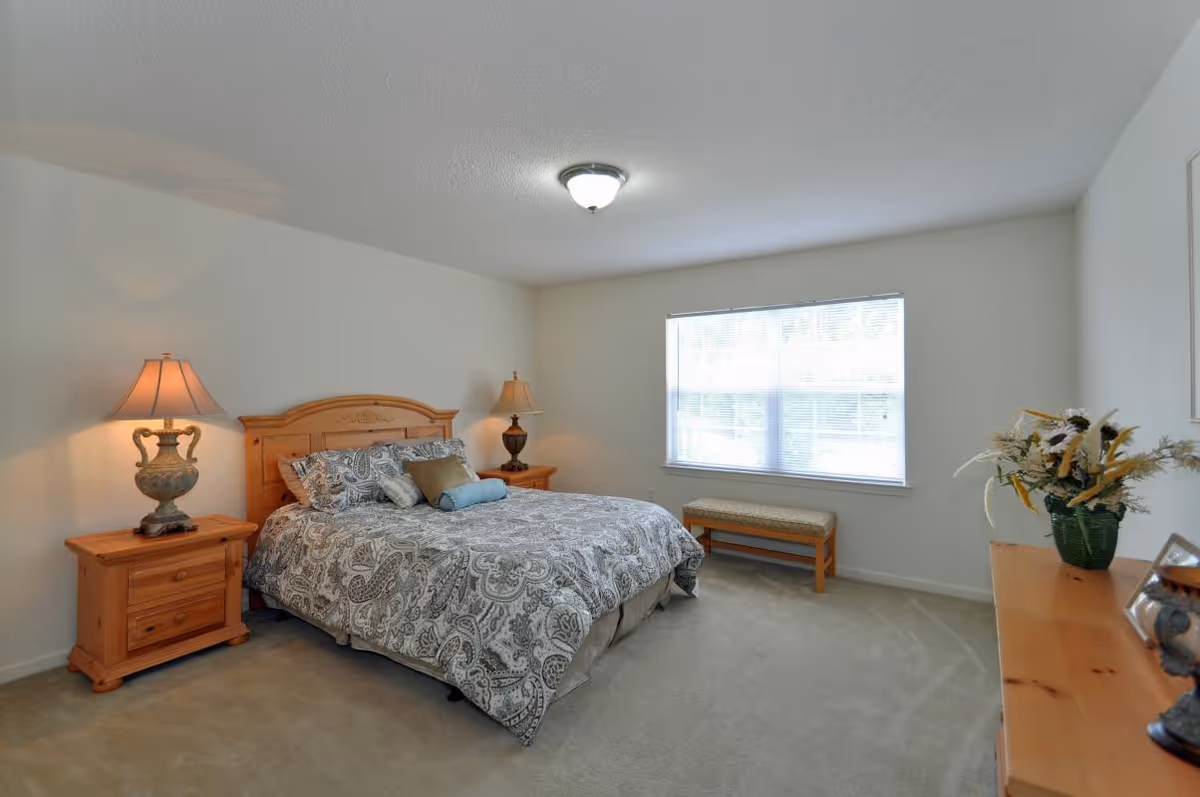 A cozy bedroom with a wooden bed frame and matching nightstands on either side. The bed is made with patterned bedding and several pillows. Two decorative table lamps are on the nightstands. A large window with blinds lets in natural light. A wooden bench is placed under the window, and a wooden dresser with a vase of flowers and decorative items is on the right side of the room. The walls are painted white and the floor is carpeted.