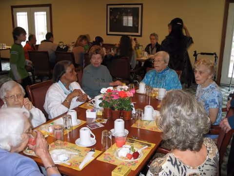 Elderly residents seated around a communal dining table with placemats, cups, and a floral centerpiece in a dining room.