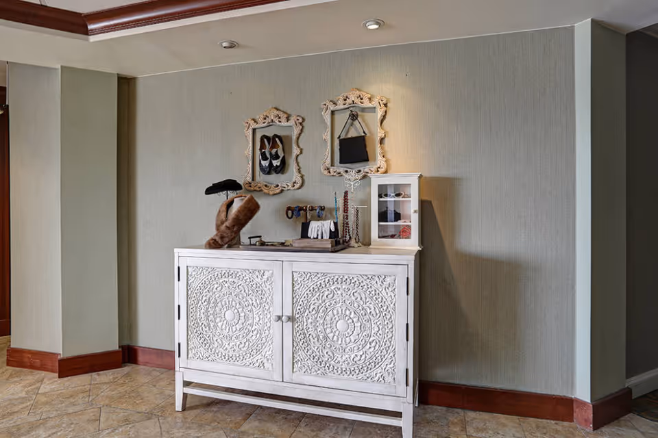 White carved console table against a pale green wall with decorative framed mirrors and jewelry displays on top.