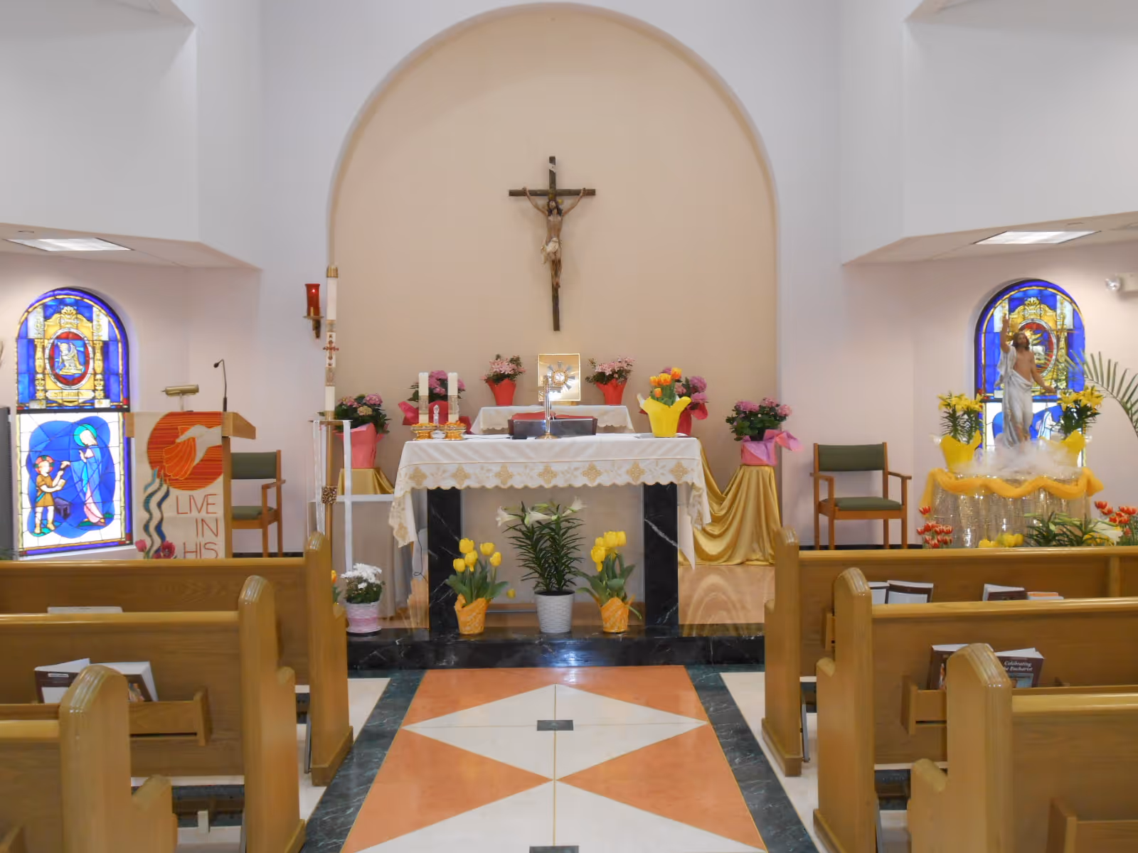 Interior view of a chapel with wooden pews, an altar adorned with flowers and a white cloth, a crucifix mounted on the wall behind the altar, stained glass windows on both sides, and religious statues and decorations.