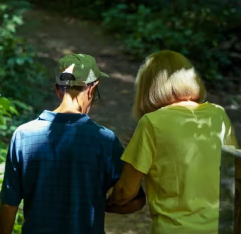 An elderly man wearing a green cap and a blue shirt walks arm-in-arm with an elderly woman in a yellow shirt along a shaded forest path.