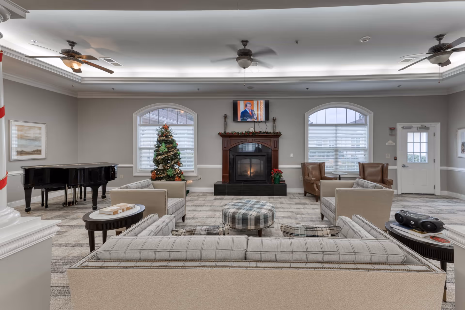 A cozy living room area in Addington Place of Johns Creek featuring a plaid sofa and matching ottoman, two beige armchairs, a fireplace with a TV mounted above it, a decorated Christmas tree, a grand piano, and two ceiling fans. The room has large windows with blinds and a door on the right side.