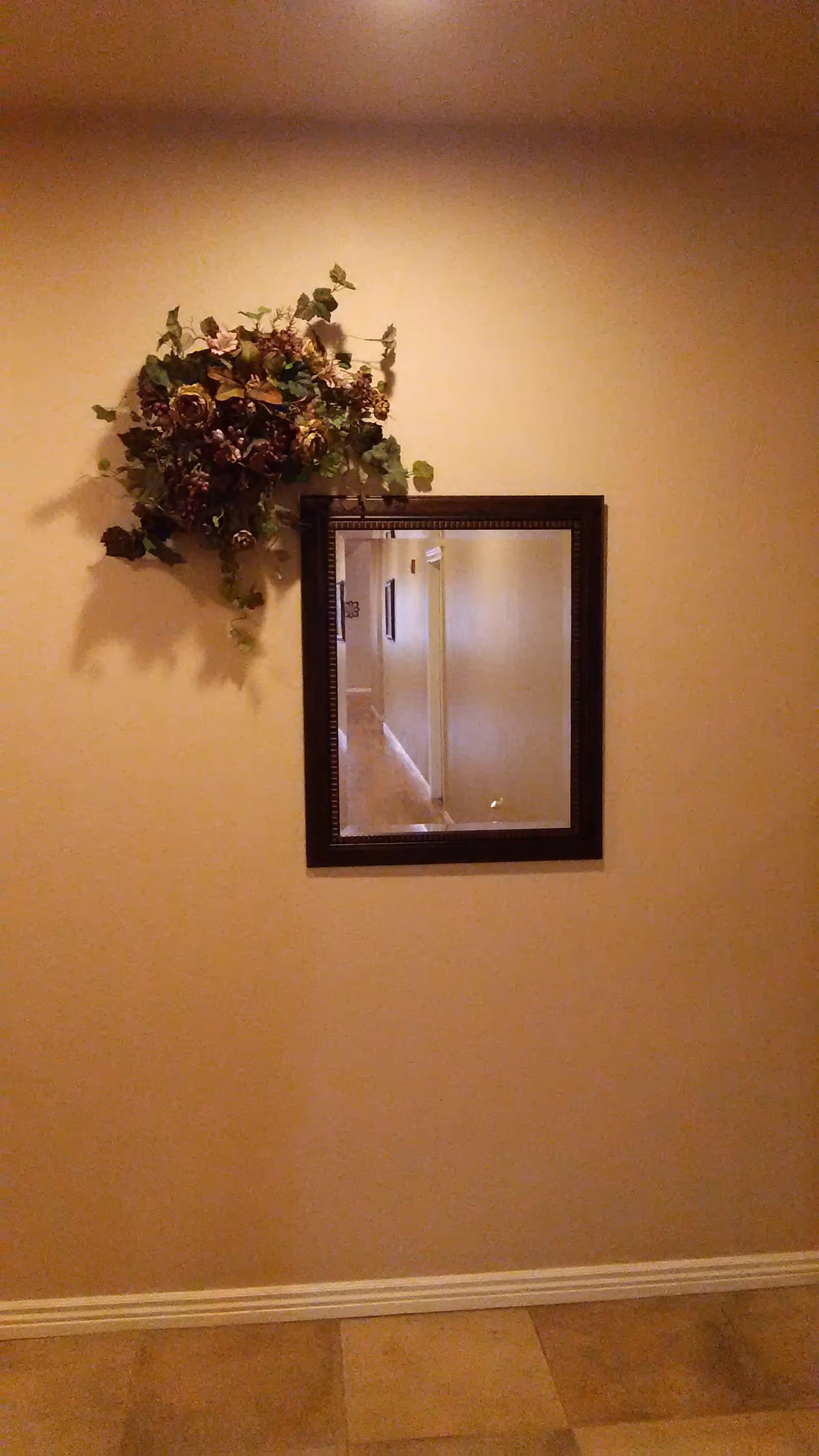 A beige wall with a decorative floral arrangement hanging above a square mirror with a dark wooden frame. The mirror reflects a hallway with beige walls and tiled flooring.