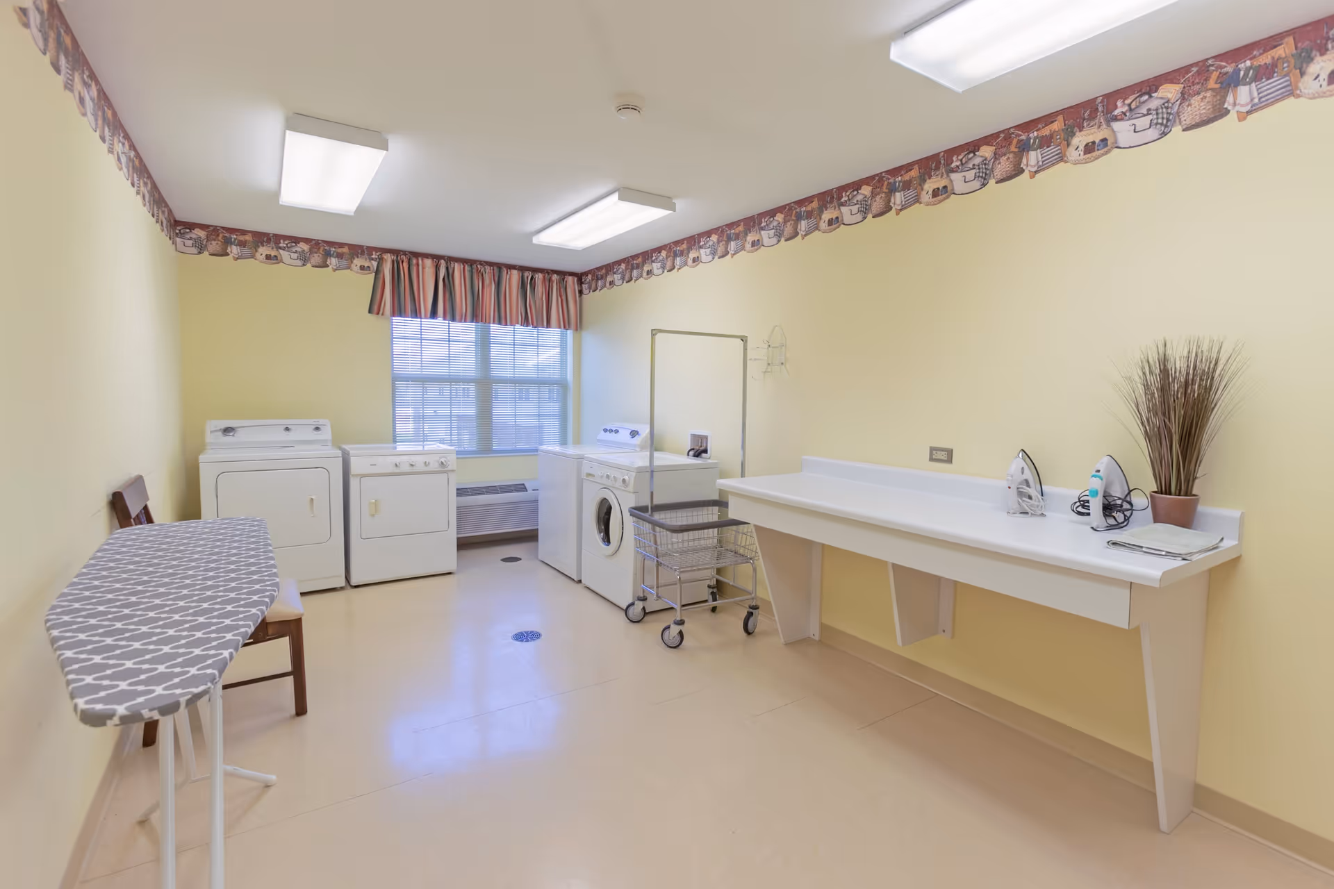 A laundry room with yellow walls and a decorative border near the ceiling. The room contains a washing machine, dryer, ironing board, and a long white counter with two irons and a potted plant. There is a window with striped curtains letting in natural light.