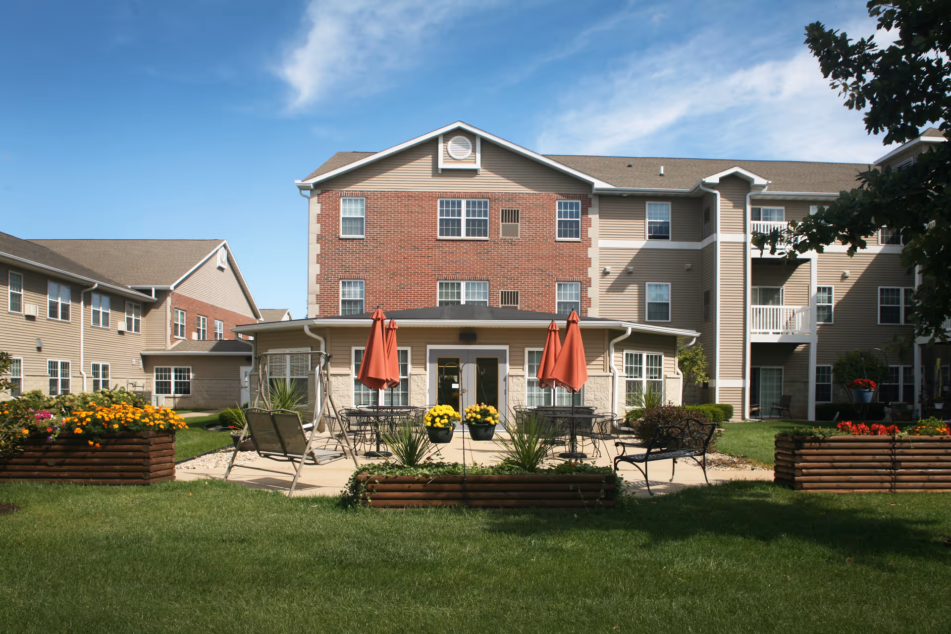 Three-story brick-and-siding senior living building with a courtyard patio, tables with red umbrellas, planters, and a green lawn.