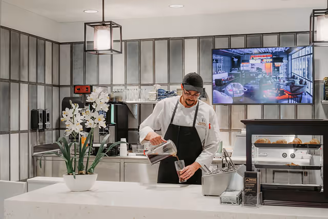 A man wearing a black apron and cap is pouring a beverage from a glass pitcher into a glass behind a white counter in a modern kitchen area. There is a white flower arrangement on the counter, and a display case with pastries to the right. A large screen mounted on the wall shows an image of a lounge area.