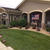 Exterior view of a senior living facility with beige walls, stone pillars, and an American flag hanging under a covered porch. There is a well-maintained lawn with small American flags placed along the sidewalk and landscaping with bushes and rocks.