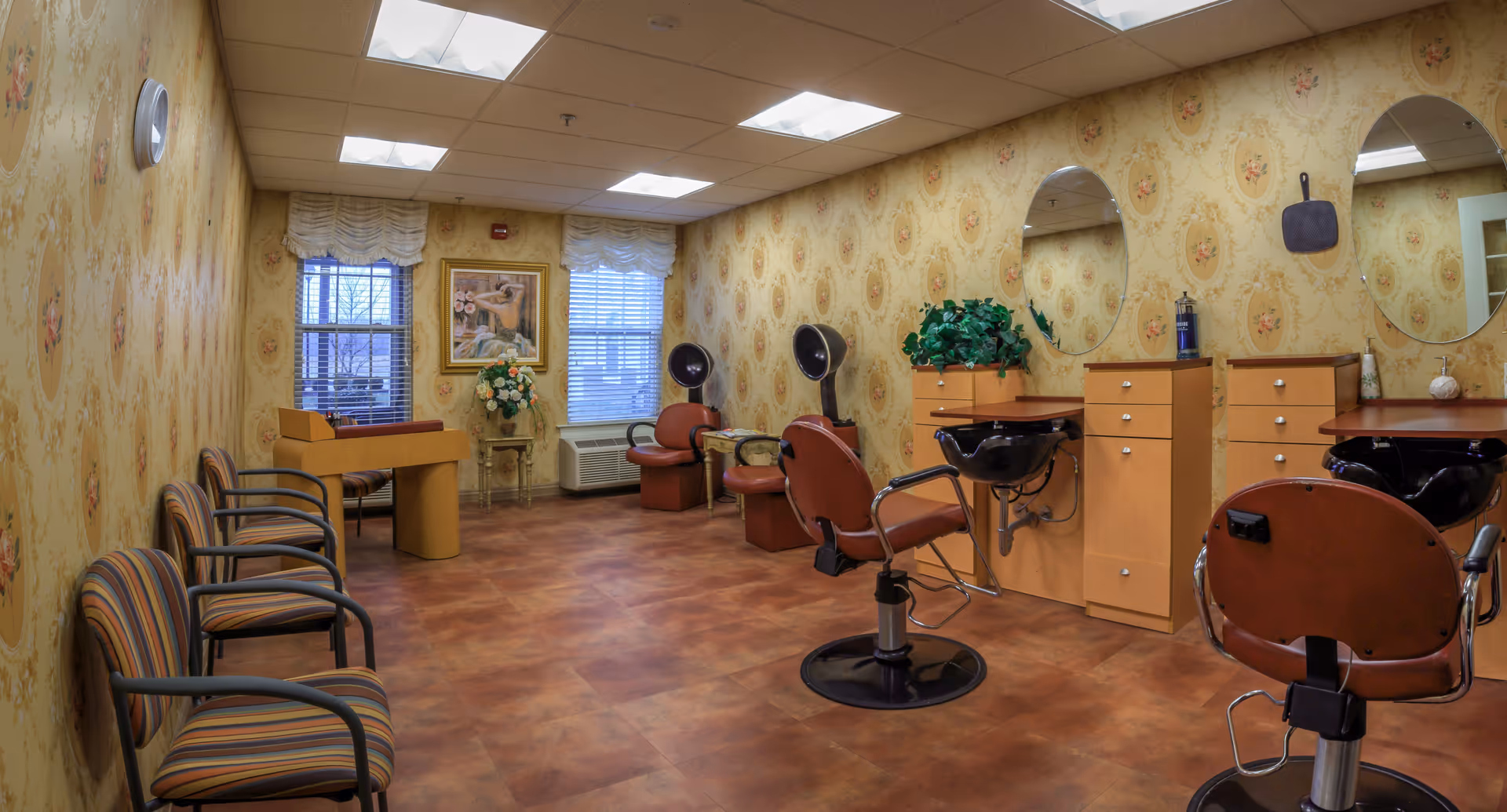 Interior view of a salon room with multiple styling chairs, hair washing stations, and mirrors on the wall. There are striped waiting chairs along one wall, floral wallpaper, two windows with blinds, a small table with flowers, and a framed painting between the windows.