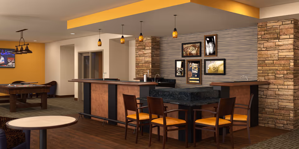 A cozy interior common area featuring a bar with four wooden chairs with orange cushions, pendant lights hanging from the ceiling, and a stone accent wall with framed pictures. In the background, there is a pool table and a wall-mounted TV, with warm yellow and beige tones throughout the space.