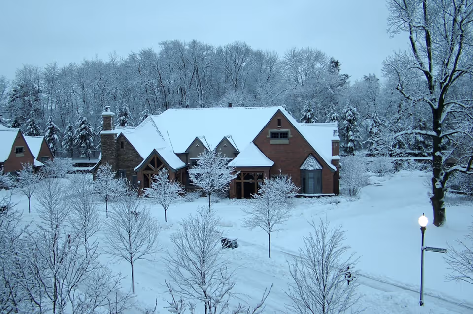 A large brick and stone building covered in snow surrounded by snow-covered trees and a snow-covered ground, with a street lamp on the right side and a forest in the background.
