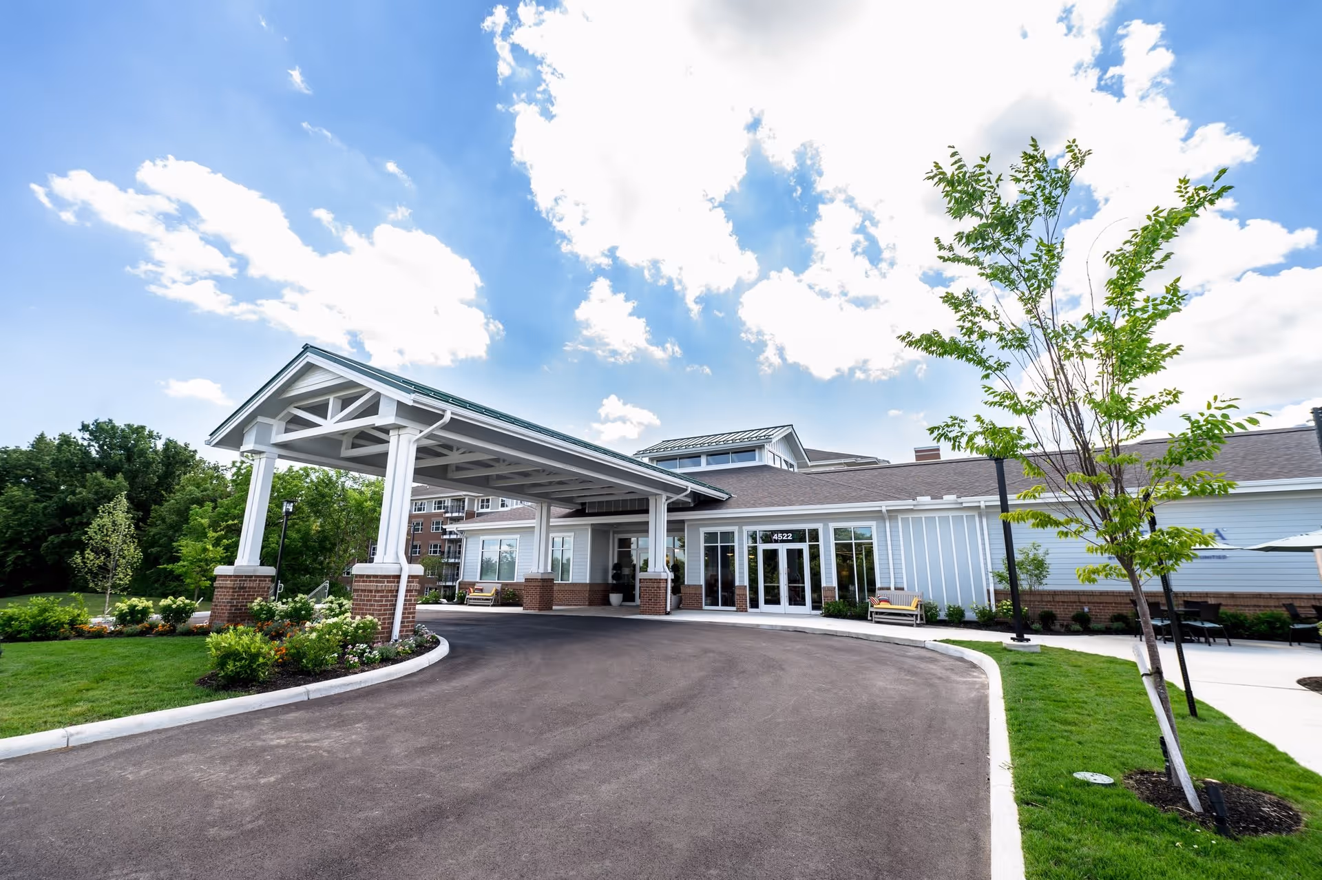 Front exterior view of Verena at Hilliard facility showing a covered entrance with white pillars and brick bases, a driveway, green lawn, trees, and a partly cloudy blue sky.