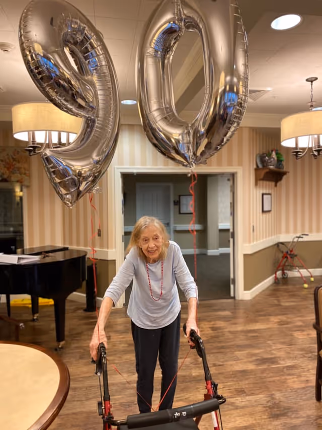 An elderly woman using a walker inside a room with wooden flooring and beige striped wallpaper. She is smiling and standing under large silver balloons shaped as the numbers 9 and 0, indicating a 90th birthday celebration. The room has a piano, round tables, chairs, and warm lighting fixtures.