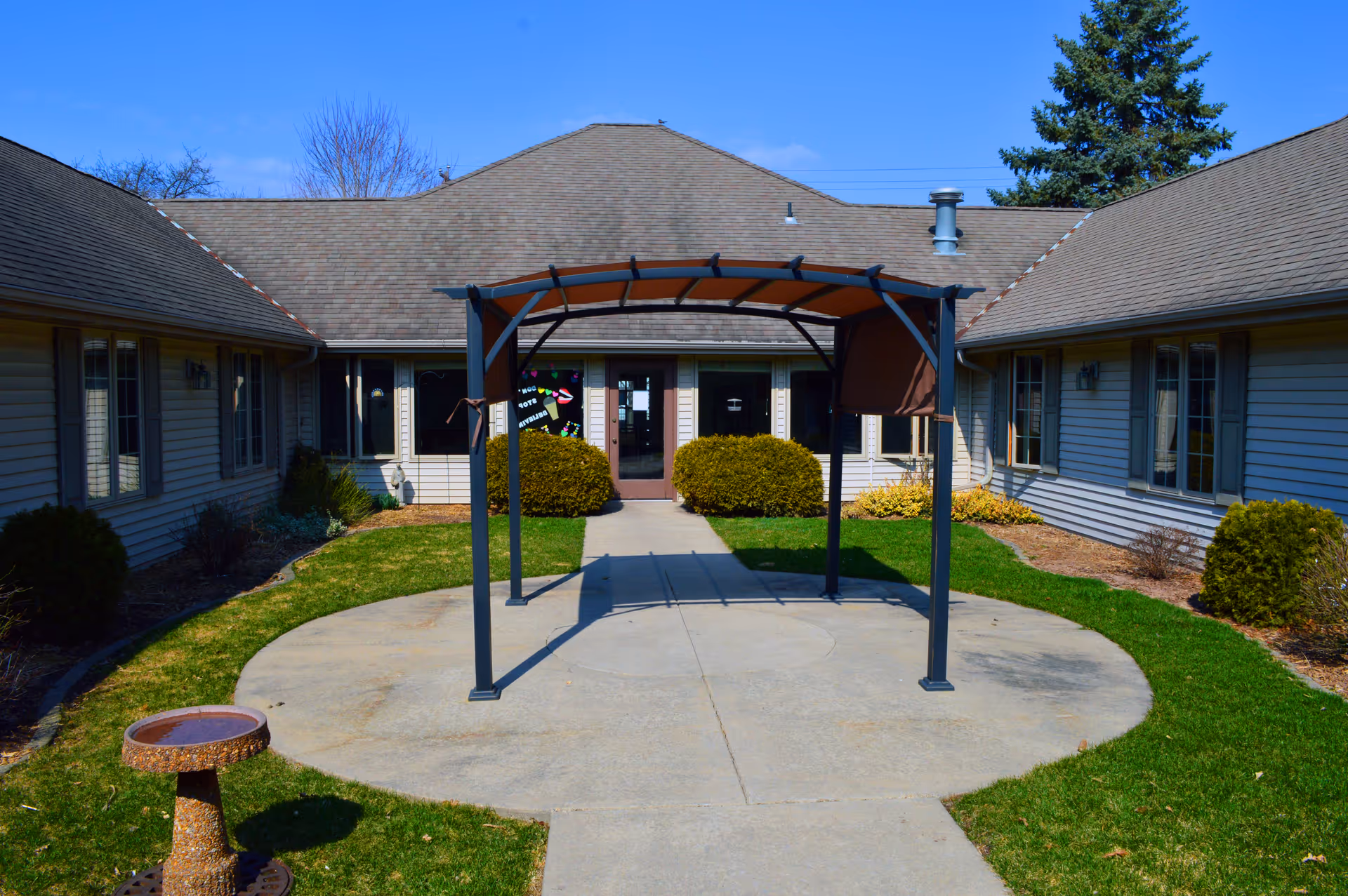 Courtyard with a metal pergola and walkway leading to the front entrance of a single-story memory care building.