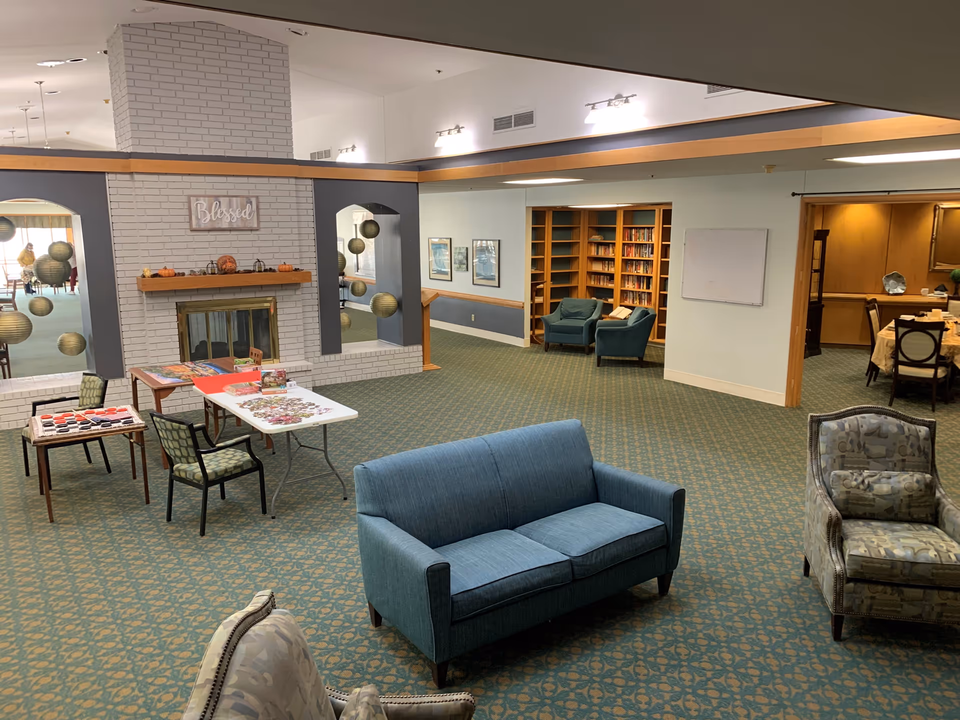 A spacious common area in an assisted living facility with a blue couch and patterned armchairs on a green carpet. There is a white brick fireplace with a wooden mantel decorated with small pumpkins and a sign that reads 'Blessed'. Two tables with chairs are set up with puzzles and games. In the background, there is a bookshelf with two blue armchairs and a dining area visible through an open doorway.