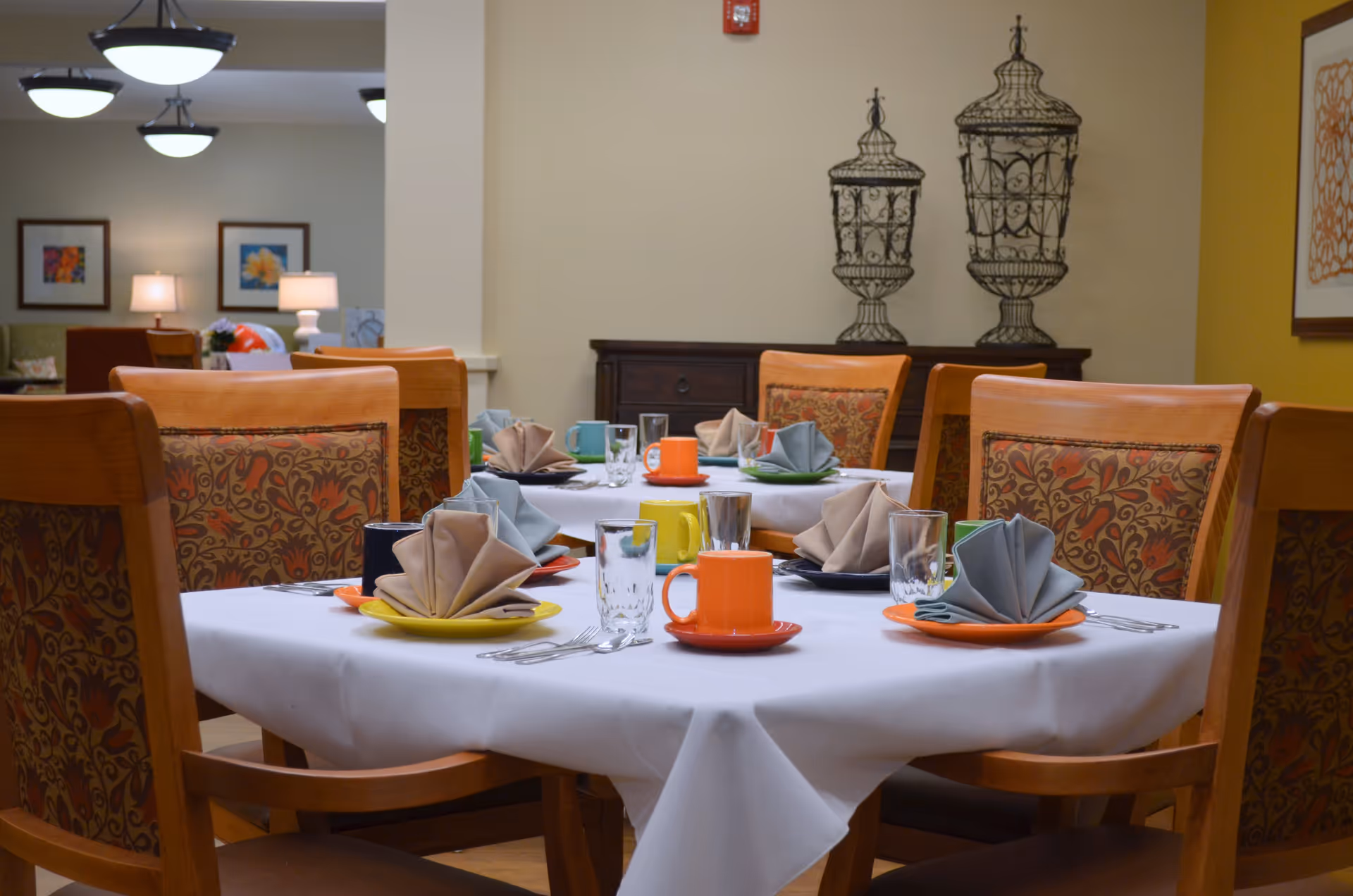 A dining room with a table set for six people. The table is covered with a white tablecloth and has colorful plates, mugs, folded napkins, and glasses. The chairs have wooden frames with patterned upholstery. In the background, there are decorative wire sculptures on a wooden sideboard and framed artwork on the walls.
