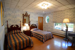 A bedroom with two twin beds featuring wooden headboards and striped bedspreads. Between the beds is a nightstand with a lamp and a decorative mirror above it. The room has patterned wallpaper, a round window with a view of greenery outside, and a table with a lamp near a larger window with a Roman shade.
