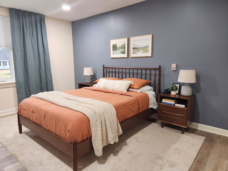 A neatly made bed with rust-colored bedding and a cream knitted throw blanket in a bedroom. The bed has a wooden frame and is flanked by two wooden nightstands, each with a lamp. Above the bed are two framed landscape paintings. The room has a blue accent wall and a window with blue curtains on the left side.